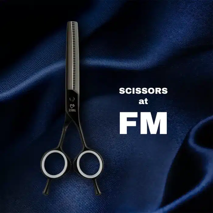 A pair of black hair thinning scissors with white inner rings is placed on dark blue fabric. The text SCISSORS at FM appears in white beside the scissors.