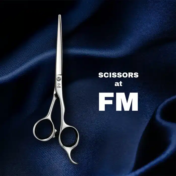 A pair of silver scissors with sleek, shiny blades displayed on a textured dark blue fabric background. The text SCISSORS at FM is written beside it in bold white letters.